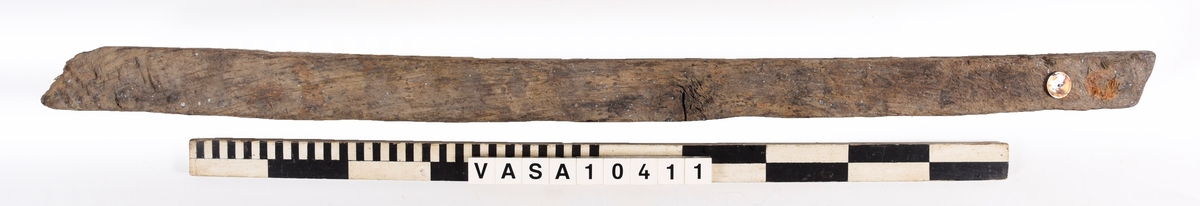 varpade timmer med en vinklade ände. 6 spikhål på ena yta, och en till möjlig på den andra sida. En kvist i mitten av föremålet, synliga på 3 ytor. 
Avsplittrad på ena ände. Mycket vittring. Torr, med djup sprickor i träfibrer. Rost fläck bredvid fyndnummer.