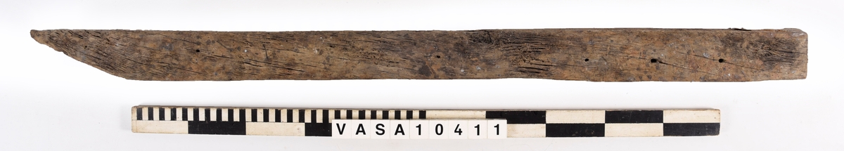 varpade timmer med en vinklade ände. 6 spikhål på ena yta, och en till möjlig på den andra sida. En kvist i mitten av föremålet, synliga på 3 ytor. 
Avsplittrad på ena ände. Mycket vittring. Torr, med djup sprickor i träfibrer. Rost fläck bredvid fyndnummer.