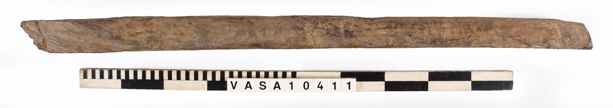 varpade timmer med en vinklade ände. 6 spikhål på ena yta, och en till möjlig på den andra sida. En kvist i mitten av föremålet, synliga på 3 ytor. 
Avsplittrad på ena ände. Mycket vittring. Torr, med djup sprickor i träfibrer. Rost fläck bredvid fyndnummer.