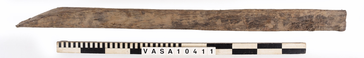 varpade timmer med en vinklade ände. 6 spikhål på ena yta, och en till möjlig på den andra sida. En kvist i mitten av föremålet, synliga på 3 ytor. 
Avsplittrad på ena ände. Mycket vittring. Torr, med djup sprickor i träfibrer. Rost fläck bredvid fyndnummer.