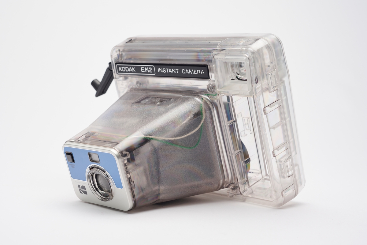Kodak EK2 Instant Camera, også kalt The Handle, i gjennomsiktig plast. Kameraet er utstyrt med bl.a. autofokus, manuell bildeutløser, mulighet for avtagbar blits og anvendte Kodaks egen PR10 instant film. Kameraet er produsert av Kodak og er en av få som har fått beholde sitt originale navnemerke, da dette kamera mest sannsynlig aldri ble solgt, men har fungert som et visningseksemplar i butikk.  
Polaroid fikk på alvor en konkurrent på markedet da Kodak begynte å produsere instant kameraer og film i 1976. På 1960-tallet hadde Kodak produsert film for Polaroid, men nå var stemningen en annen. Dette var ikke et samarbeid og Polaroid, som hadde patent på produktet, svarte med å gå til rettssak. Det var en langvarig rettssak. Kodak ble nødt til å trekke sine instant-produkter fra markedet i 1986. I 1990 var erstatningssummen avklart og året etter måtte Kodak betale Polaroid 925 millioner amerikanske dollar (inkludert renter). Dette var den største erstatningssummen noen gang frem til 2012. I tillegg måtte Kodak kompensere sine kunder som hadde kjøpt instant kameraer av dem fra 1976 til 1986, da de nå ikke lenger fikk kjøpt film som passet. Kunden ble bedt om å ringe et gratisnummer og registrere seg for å få tilsendt en pakke i posten, som inneholdt instrukser for hvordan de kunne få kompensasjon. Ofte involverte dette at de måtte ta av kameraets navnemerke og sende tilbake som et bevis for at de eide kameraet. I dag er instant kameraer fra Kodak uten navnemerket mer vanlig enn med.