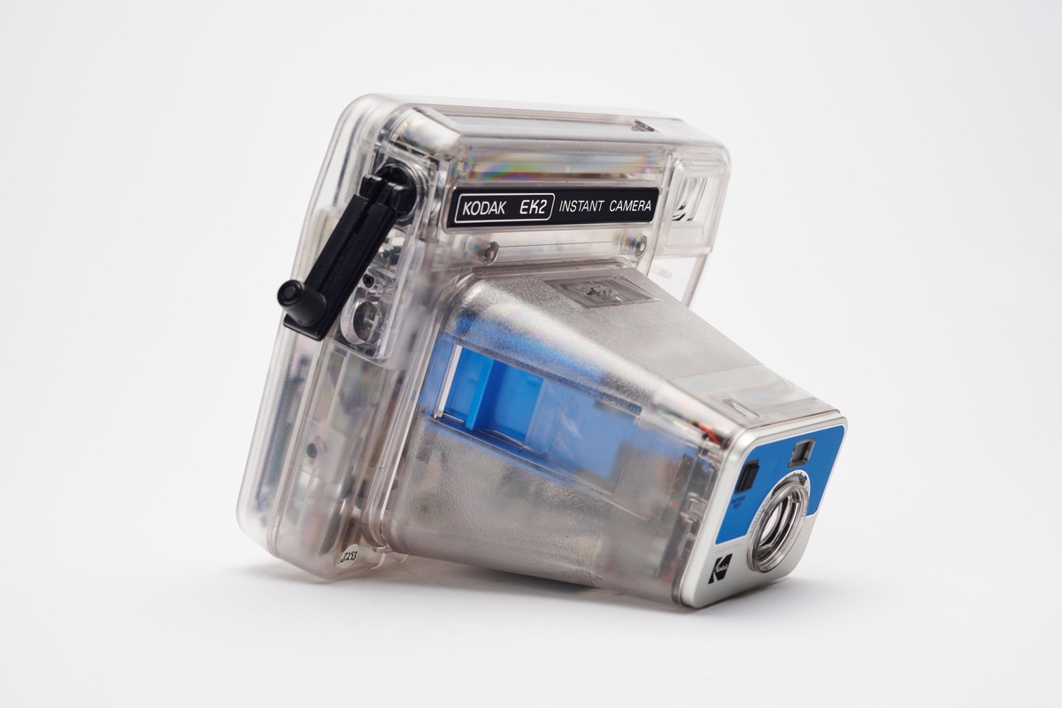 Kodak EK2 Instant Camera, også kalt The Handle, i gjennomsiktig plast. Kameraet er utstyrt med bl.a. autofokus, manuell bildeutløser, mulighet for avtagbar blits og anvendte Kodaks egen PR10 instant film. Kameraet er produsert av Kodak og er en av få som har fått beholde sitt originale navnemerke, da dette kamera mest sannsynlig aldri ble solgt, men har fungert som et visningseksemplar i butikk.  
Polaroid fikk på alvor en konkurrent på markedet da Kodak begynte å produsere instant kameraer og film i 1976. På 1960-tallet hadde Kodak produsert film for Polaroid, men nå var stemningen en annen. Dette var ikke et samarbeid og Polaroid, som hadde patent på produktet, svarte med å gå til rettssak. Det var en langvarig rettssak. Kodak ble nødt til å trekke sine instant-produkter fra markedet i 1986. I 1990 var erstatningssummen avklart og året etter måtte Kodak betale Polaroid 925 millioner amerikanske dollar (inkludert renter). Dette var den største erstatningssummen noen gang frem til 2012. I tillegg måtte Kodak kompensere sine kunder som hadde kjøpt instant kameraer av dem fra 1976 til 1986, da de nå ikke lenger fikk kjøpt film som passet. Kunden ble bedt om å ringe et gratisnummer og registrere seg for å få tilsendt en pakke i posten, som inneholdt instrukser for hvordan de kunne få kompensasjon. Ofte involverte dette at de måtte ta av kameraets navnemerke og sende tilbake som et bevis for at de eide kameraet. I dag er instant kameraer fra Kodak uten navnemerket mer vanlig enn med.
