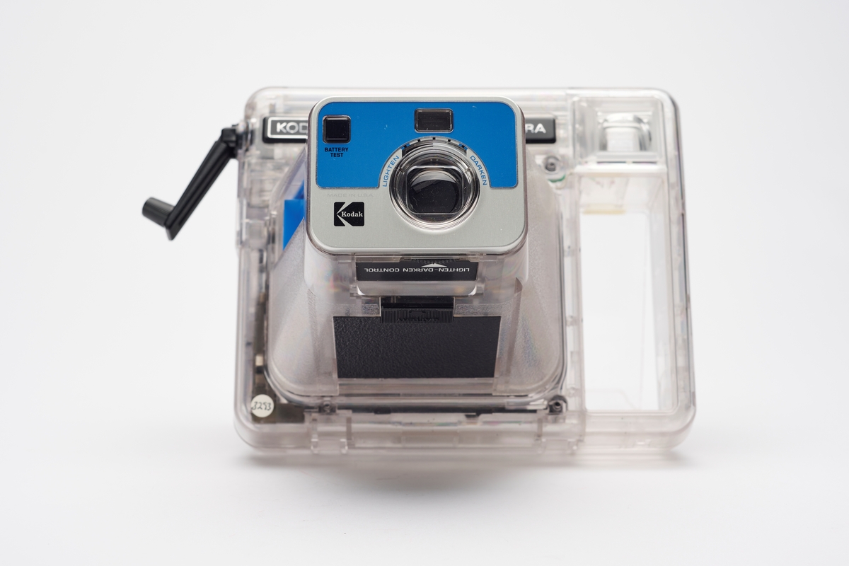 Kodak EK2 Instant Camera, også kalt The Handle, i gjennomsiktig plast. Kameraet er utstyrt med bl.a. autofokus, manuell bildeutløser, mulighet for avtagbar blits og anvendte Kodaks egen PR10 instant film. Kameraet er produsert av Kodak og er en av få som har fått beholde sitt originale navnemerke, da dette kamera mest sannsynlig aldri ble solgt, men har fungert som et visningseksemplar i butikk.  
Polaroid fikk på alvor en konkurrent på markedet da Kodak begynte å produsere instant kameraer og film i 1976. På 1960-tallet hadde Kodak produsert film for Polaroid, men nå var stemningen en annen. Dette var ikke et samarbeid og Polaroid, som hadde patent på produktet, svarte med å gå til rettssak. Det var en langvarig rettssak. Kodak ble nødt til å trekke sine instant-produkter fra markedet i 1986. I 1990 var erstatningssummen avklart og året etter måtte Kodak betale Polaroid 925 millioner amerikanske dollar (inkludert renter). Dette var den største erstatningssummen noen gang frem til 2012. I tillegg måtte Kodak kompensere sine kunder som hadde kjøpt instant kameraer av dem fra 1976 til 1986, da de nå ikke lenger fikk kjøpt film som passet. Kunden ble bedt om å ringe et gratisnummer og registrere seg for å få tilsendt en pakke i posten, som inneholdt instrukser for hvordan de kunne få kompensasjon. Ofte involverte dette at de måtte ta av kameraets navnemerke og sende tilbake som et bevis for at de eide kameraet. I dag er instant kameraer fra Kodak uten navnemerket mer vanlig enn med.