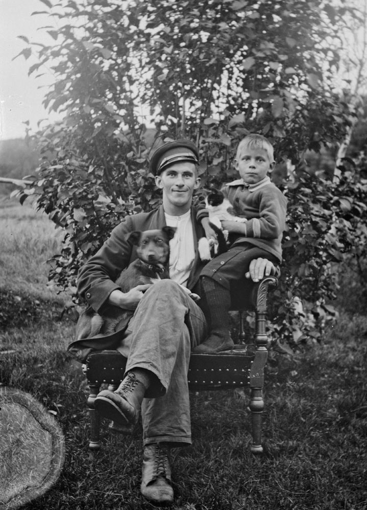 Leirfjord. Ung mann fotografert i stol ute, med gutt på armen. Gutten har svartlugger på føttene. De har en hund og en katt på fanget. Navnene er ukjent.
Stolen og katten er også på bilde LEF.F.02246