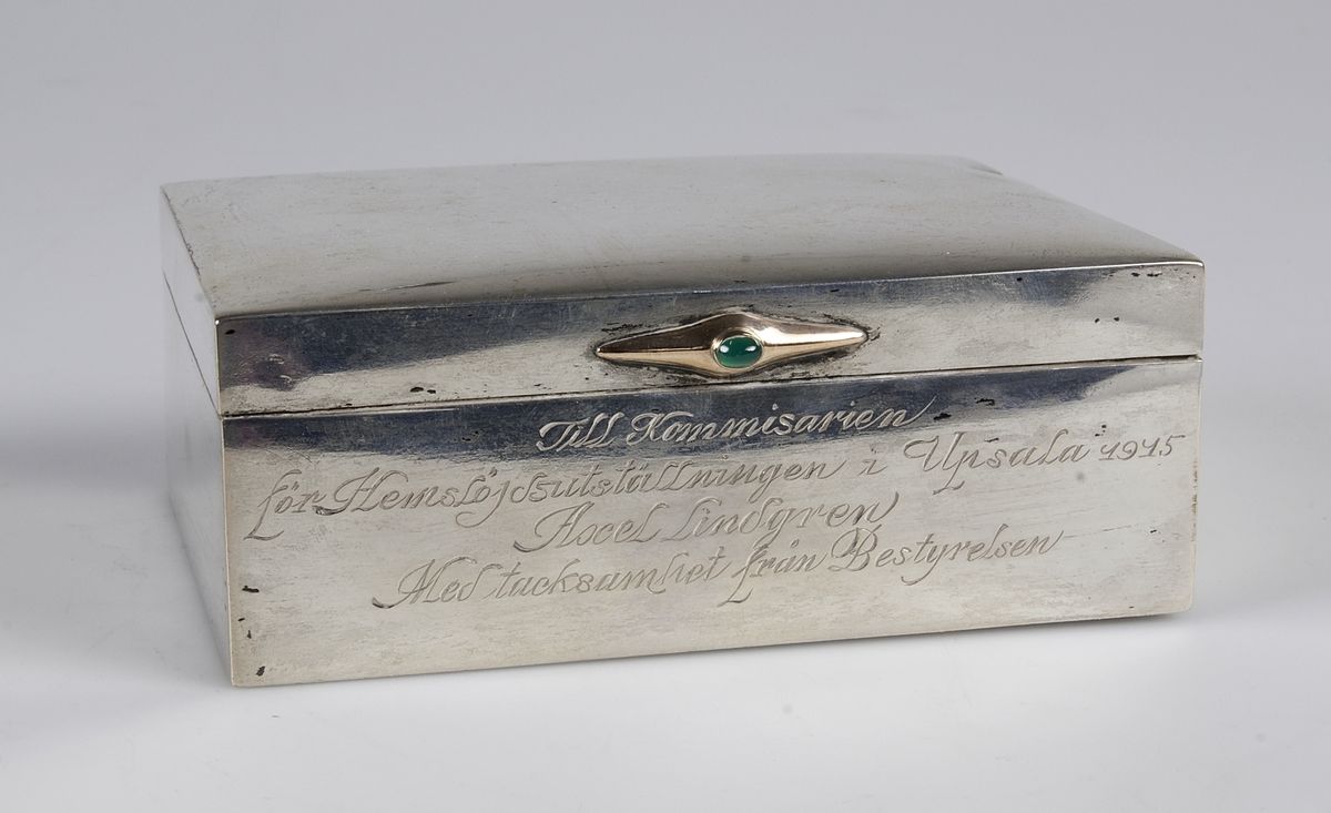Av silver med beslag av guld med grön sten. på framsidan ingraverat: "Till kommissarie för hemslöjdsutställningen i Uppsala 1914 Axel Lindgren. Med tacksamhet från bestyrelsen".
Invändigt träfodrad. Blybotten. Stämplar på lockets kant: KG M G AB, U, tre kronor, S, otydbar stämpel, L7.