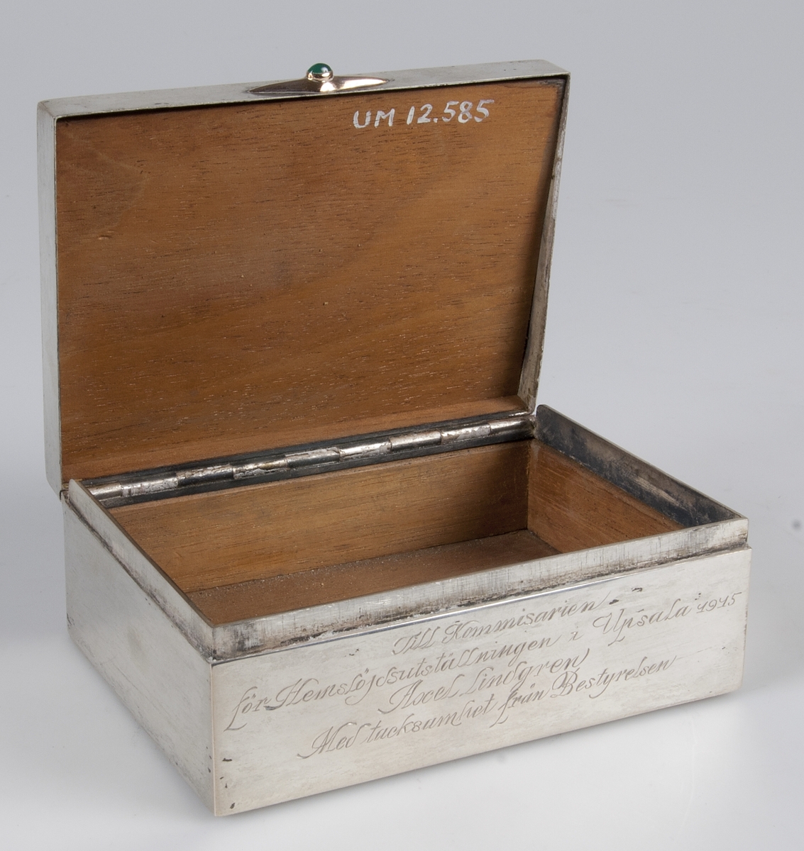 Av silver med beslag av guld med grön sten. på framsidan ingraverat: "Till kommissarie för hemslöjdsutställningen i Uppsala 1914 Axel Lindgren. Med tacksamhet från bestyrelsen".
Invändigt träfodrad. Blybotten. Stämplar på lockets kant: KG M G AB, U, tre kronor, S, otydbar stämpel, L7.