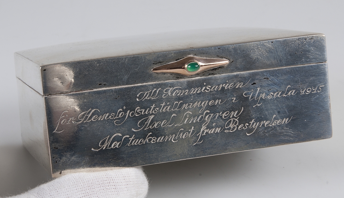 Av silver med beslag av guld med grön sten. på framsidan ingraverat: "Till kommissarie för hemslöjdsutställningen i Uppsala 1914 Axel Lindgren. Med tacksamhet från bestyrelsen".
Invändigt träfodrad. Blybotten. Stämplar på lockets kant: KG M G AB, U, tre kronor, S, otydbar stämpel, L7.