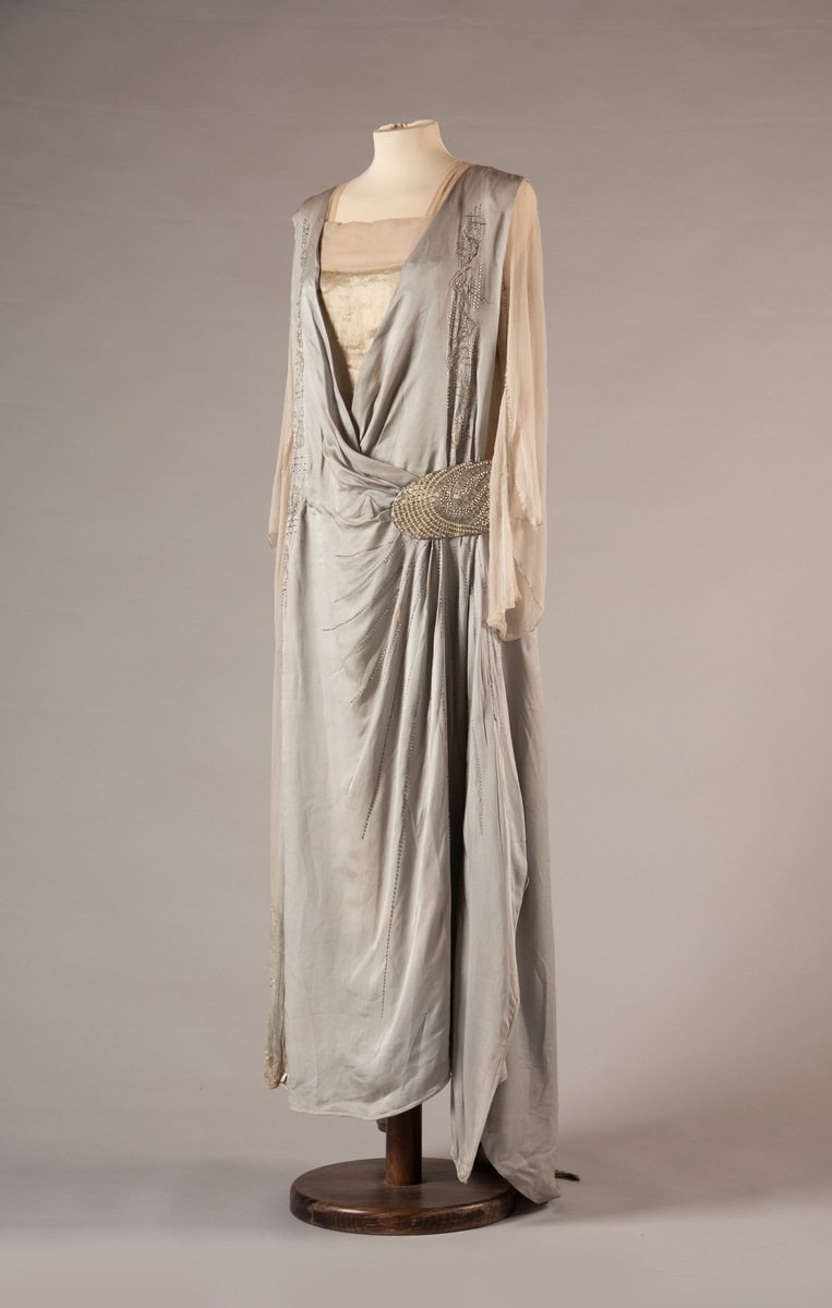 Selskapskjole i grått silkestoff med hvitt fôr i silkestoff. Kjolen er dekorert med glassperler (strass). Kjolen er produsert av Paulsson & Co i Oslo, trolig på 1920-tallet.