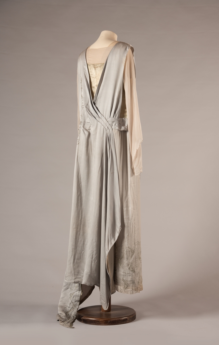 Selskapskjole i grått silkestoff med hvitt fôr i silkestoff. Kjolen er dekorert med glassperler (strass). Kjolen er produsert av Paulsson & Co i Oslo, trolig på 1920-tallet.