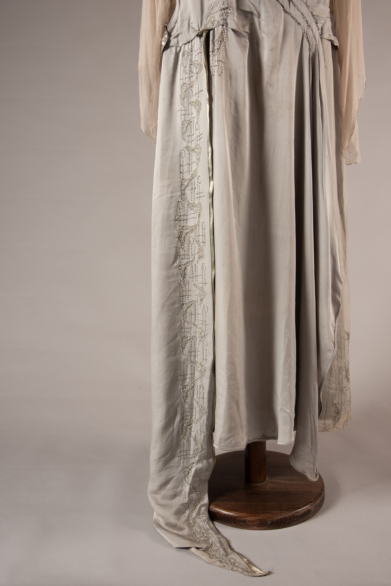 Selskapskjole i grått silkestoff med hvitt fôr i silkestoff. Kjolen er dekorert med glassperler (strass). Kjolen er produsert av Paulsson & Co i Oslo, trolig på 1920-tallet.