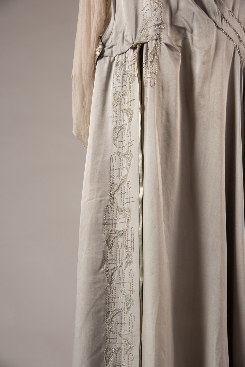 Selskapskjole i grått silkestoff med hvitt fôr i silkestoff. Kjolen er dekorert med glassperler (strass). Kjolen er produsert av Paulsson & Co i Oslo, trolig på 1920-tallet.