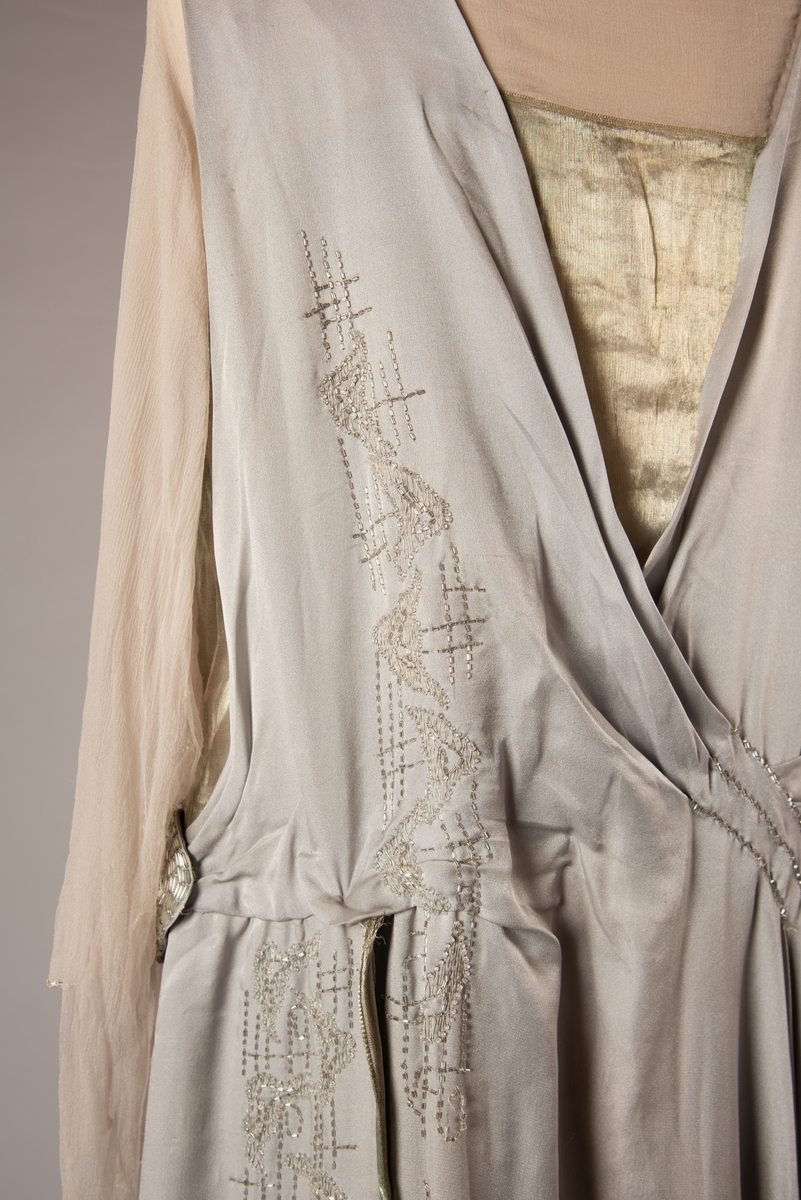 Selskapskjole i grått silkestoff med hvitt fôr i silkestoff. Kjolen er dekorert med glassperler (strass). Kjolen er produsert av Paulsson & Co i Oslo, trolig på 1920-tallet.