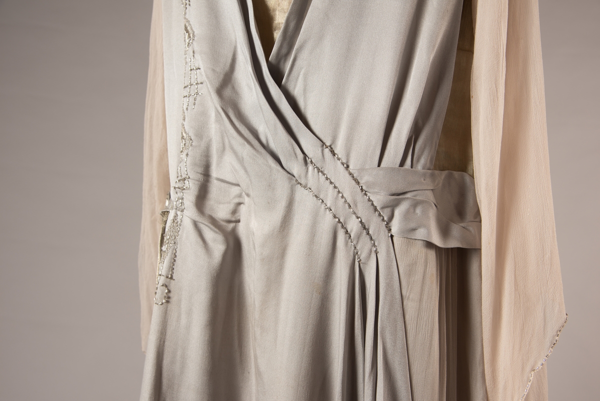 Selskapskjole i grått silkestoff med hvitt fôr i silkestoff. Kjolen er dekorert med glassperler (strass). Kjolen er produsert av Paulsson & Co i Oslo, trolig på 1920-tallet.