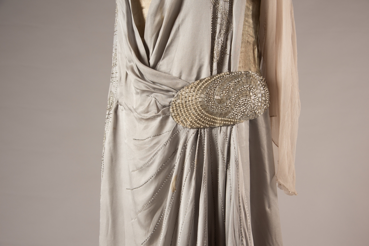 Selskapskjole i grått silkestoff med hvitt fôr i silkestoff. Kjolen er dekorert med glassperler (strass). Kjolen er produsert av Paulsson & Co i Oslo, trolig på 1920-tallet.