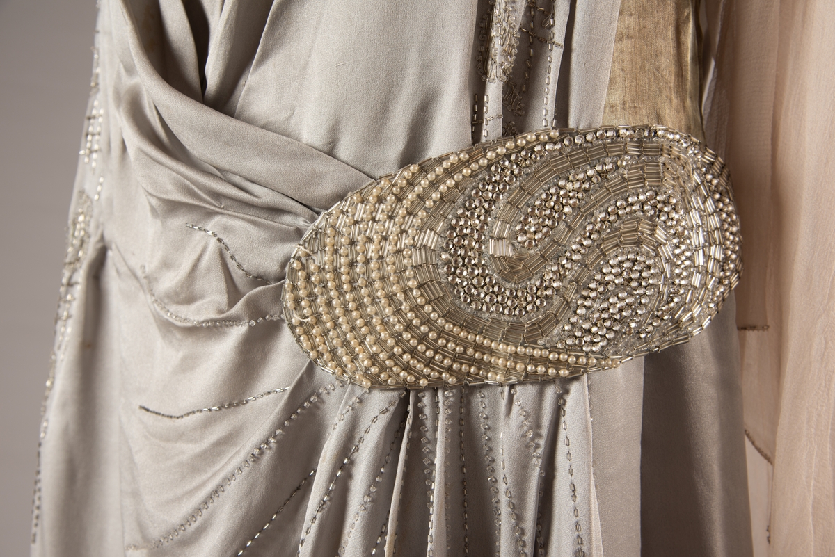 Selskapskjole i grått silkestoff med hvitt fôr i silkestoff. Kjolen er dekorert med glassperler (strass). Kjolen er produsert av Paulsson & Co i Oslo, trolig på 1920-tallet.