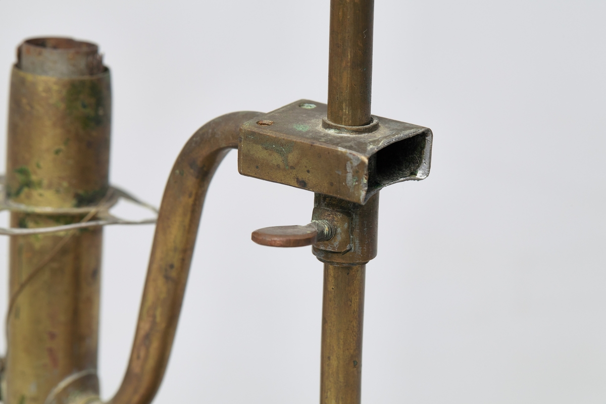 Bordlampe av messing med innhul fot. I foten står en stang med bærehempe i toppen. Oljebeholder og lampe sitter på hver sin side av stangen, og er festet til denne med en glideanordning som kan skrues fast i ønskelig høyde. Lampeglasset er borte.