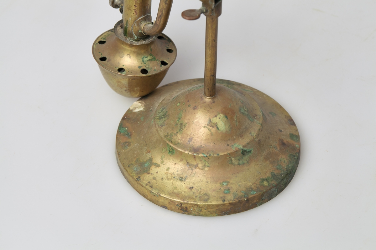 Bordlampe av messing med innhul fot. I foten står en stang med bærehempe i toppen. Oljebeholder og lampe sitter på hver sin side av stangen, og er festet til denne med en glideanordning som kan skrues fast i ønskelig høyde. Lampeglasset er borte.