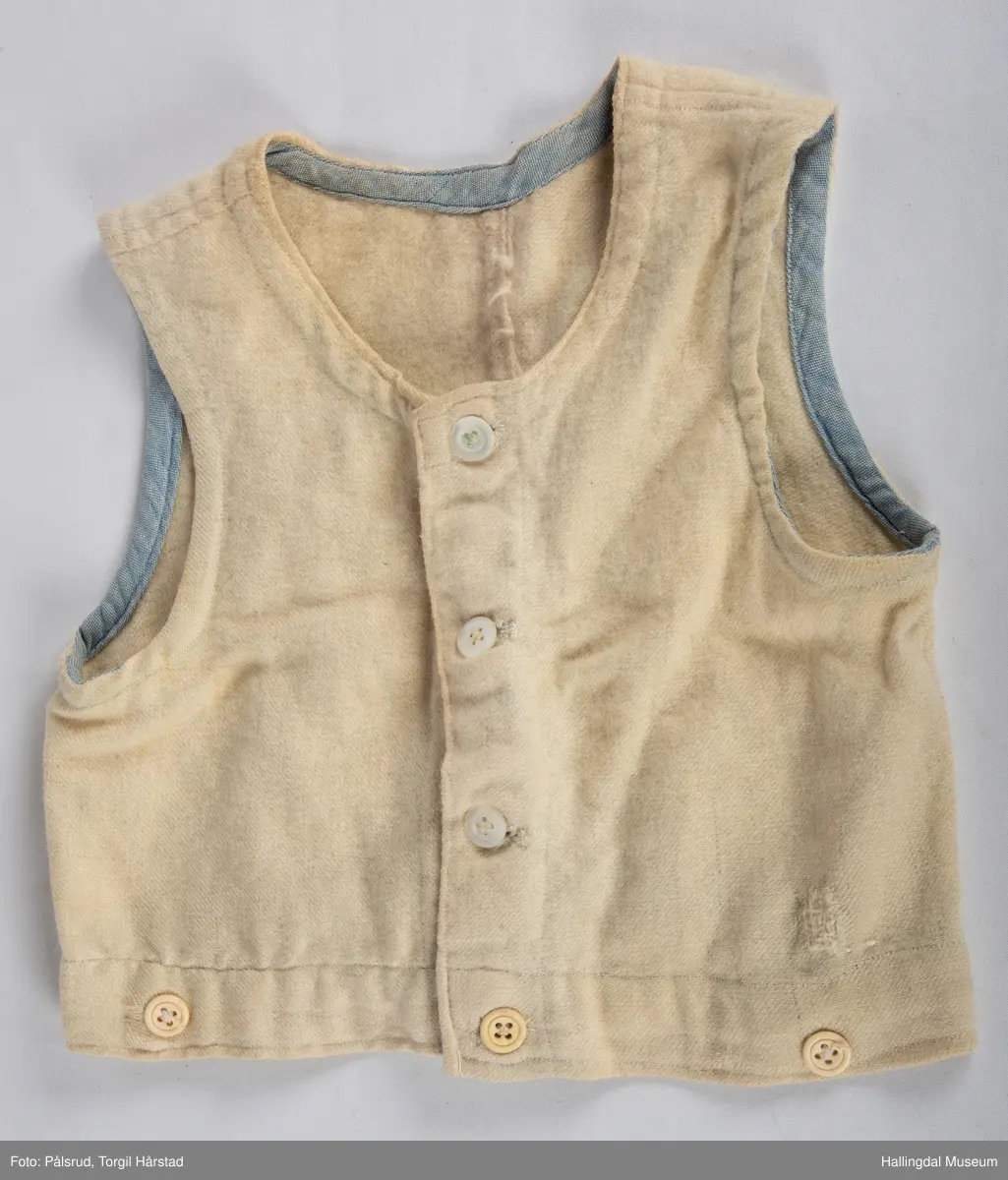 Vest i hvitt vevd ullstoff med påsydd blått skråbånd på innsiden av hals- og ermekant. Tre perlemorsknapper i front i tillegg til tre gule (plast?) knapper på nedre kant.