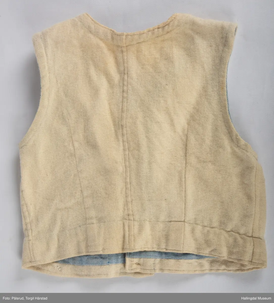 Vest i hvitt vevd ullstoff med påsydd blått skråbånd på innsiden av hals- og ermekant. Tre perlemorsknapper i front i tillegg til tre gule (plast?) knapper på nedre kant.