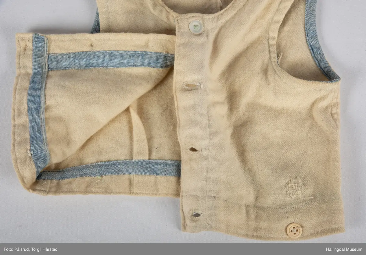 Vest i hvitt vevd ullstoff med påsydd blått skråbånd på innsiden av hals- og ermekant. Tre perlemorsknapper i front i tillegg til tre gule (plast?) knapper på nedre kant.
