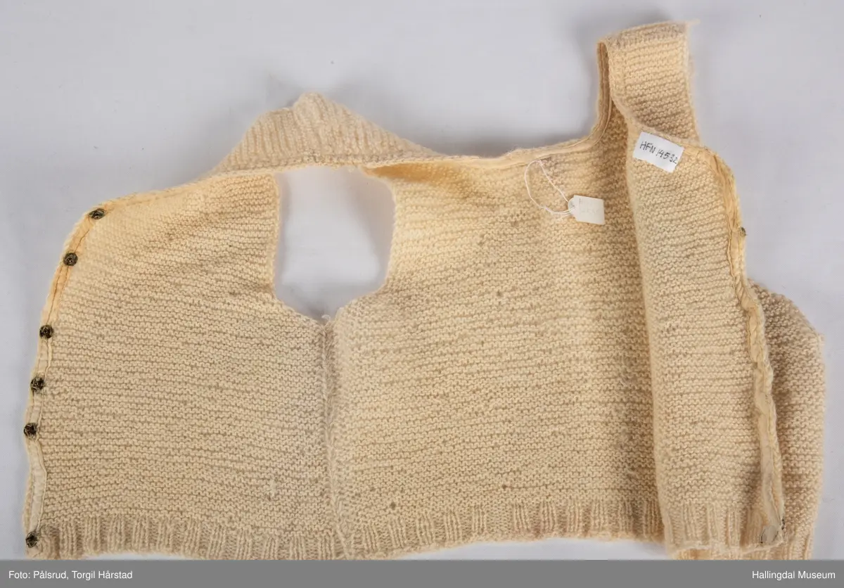 Strikket vest (trøye) i hvit ull. Hvitt bomulls-skråbånd rundt halskant og nedover åpning. Åpningen forran lukkes med 6 trykknapper i metall.