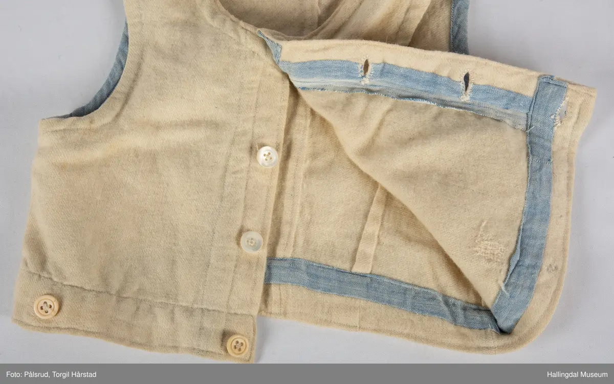 Vest i hvitt vevd ullstoff med påsydd blått skråbånd på innsiden av hals- og ermekant. Tre perlemorsknapper i front i tillegg til tre gule (plast?) knapper på nedre kant.