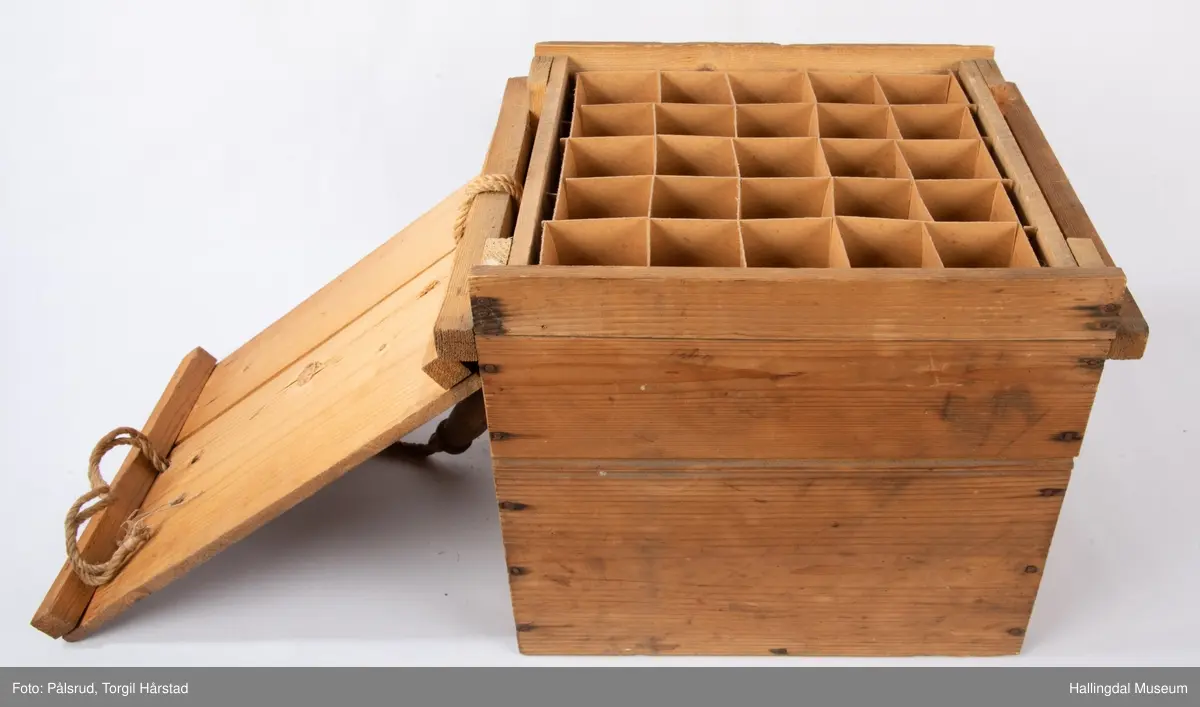 Emballasje til frakt av egg. Kassen er i ubehandlet tre, har et lokk med et dreid håndtak festet med tau. Inneholder flere seksjoner inndelt med papp der hver seksjon kunne oppbevare 25 egg. En bruksanvisning i papp ligger på toppen.