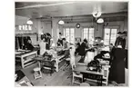 Beklädnadsateljéerna på NK (Nordiska Kompaniet). I lokalen arbetar både män och kvinnor med sömnadsarbeten vid långa bord. I förgrunden står två symaskiner.