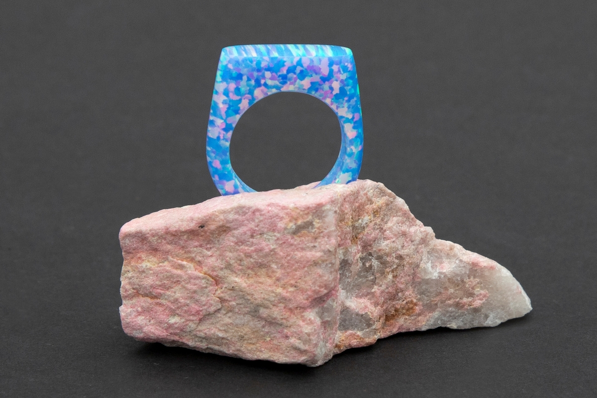 Lys blå ring med regnbue eller glittereffekter på rosa steinpodium. Ringen er lagd av syntetsik opal (bello) og podieet er av thulitt.