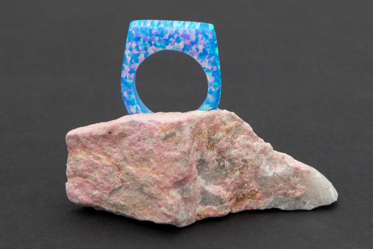 Lys blå ring med regnbue eller glittereffekter på rosa steinpodium. Ringen er lagd av syntetsik opal (bello) og podieet er av thulitt.