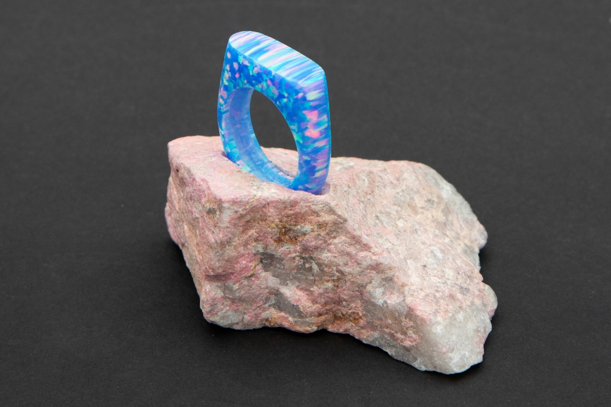 Lys blå ring med regnbue eller glittereffekter på rosa steinpodium. Ringen er lagd av syntetsik opal (bello) og podieet er av thulitt.