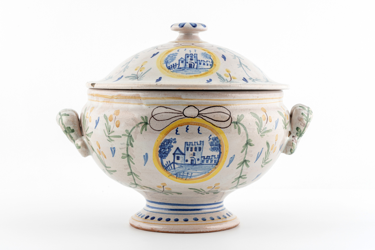 Rund terrin på stett. I keramikk, med lokk og to hanker. Beige glasur i bunn, påmalt dekor av blomster, ranker og sløyfer. Motiv av borg på ene siden, motiv av vindmølle på den andre. Disse to motivene går igjen i dekoren på lokket. Mye krakelering i glasuren inni. Har tilhørt Sara Fabricius.