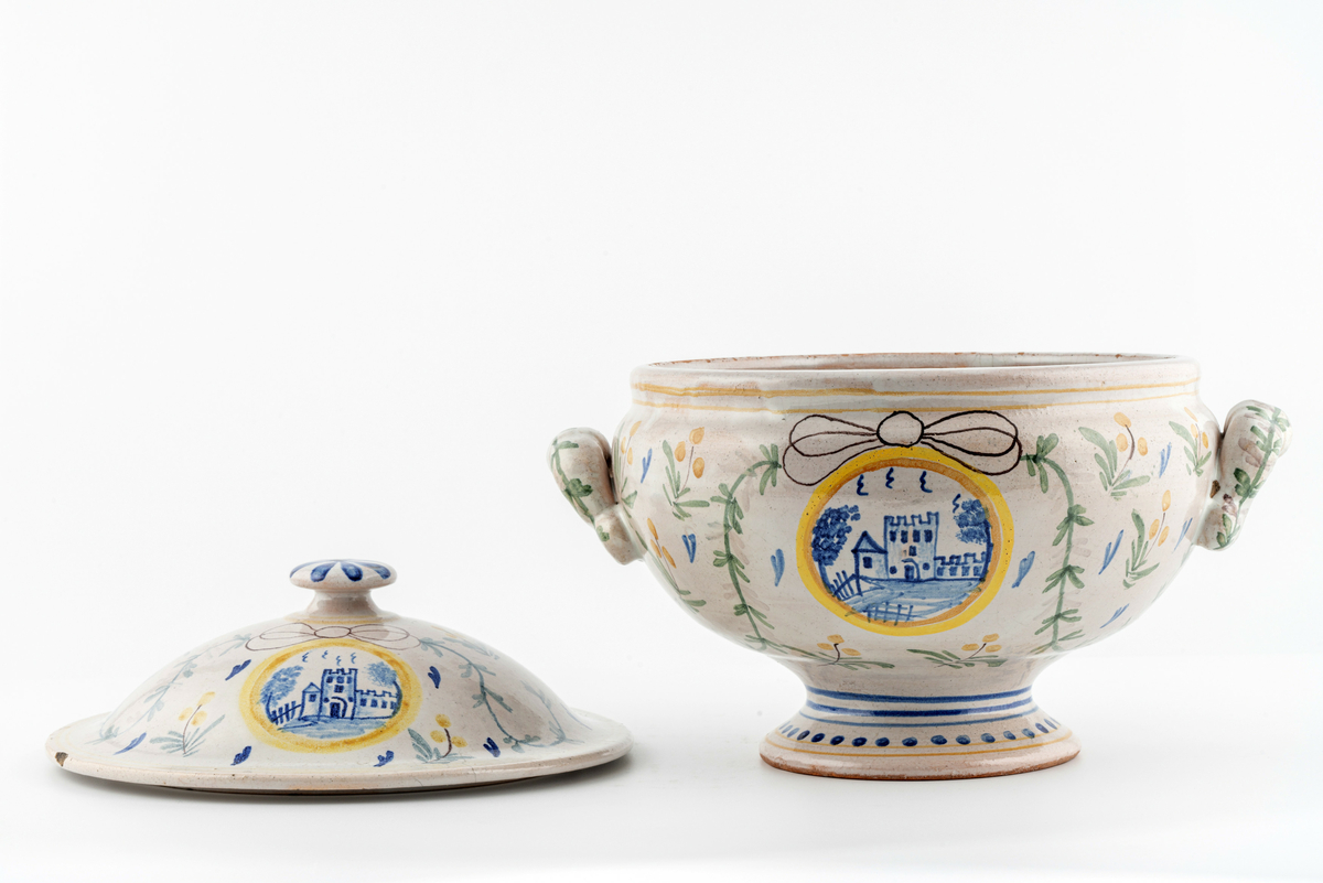 Rund terrin på stett. I keramikk, med lokk og to hanker. Beige glasur i bunn, påmalt dekor av blomster, ranker og sløyfer. Motiv av borg på ene siden, motiv av vindmølle på den andre. Disse to motivene går igjen i dekoren på lokket. Mye krakelering i glasuren inni. Har tilhørt Sara Fabricius.