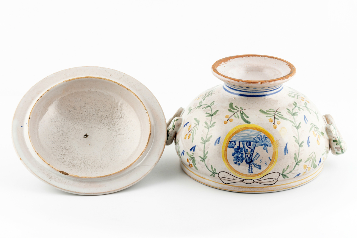 Rund terrin på stett. I keramikk, med lokk og to hanker. Beige glasur i bunn, påmalt dekor av blomster, ranker og sløyfer. Motiv av borg på ene siden, motiv av vindmølle på den andre. Disse to motivene går igjen i dekoren på lokket. Mye krakelering i glasuren inni. Har tilhørt Sara Fabricius.