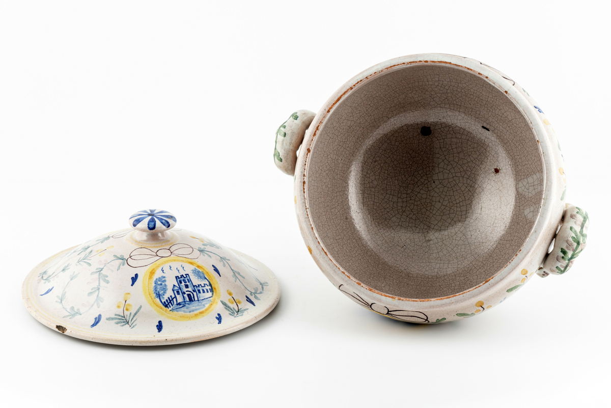 Rund terrin på stett. I keramikk, med lokk og to hanker. Beige glasur i bunn, påmalt dekor av blomster, ranker og sløyfer. Motiv av borg på ene siden, motiv av vindmølle på den andre. Disse to motivene går igjen i dekoren på lokket. Mye krakelering i glasuren inni. Har tilhørt Sara Fabricius.