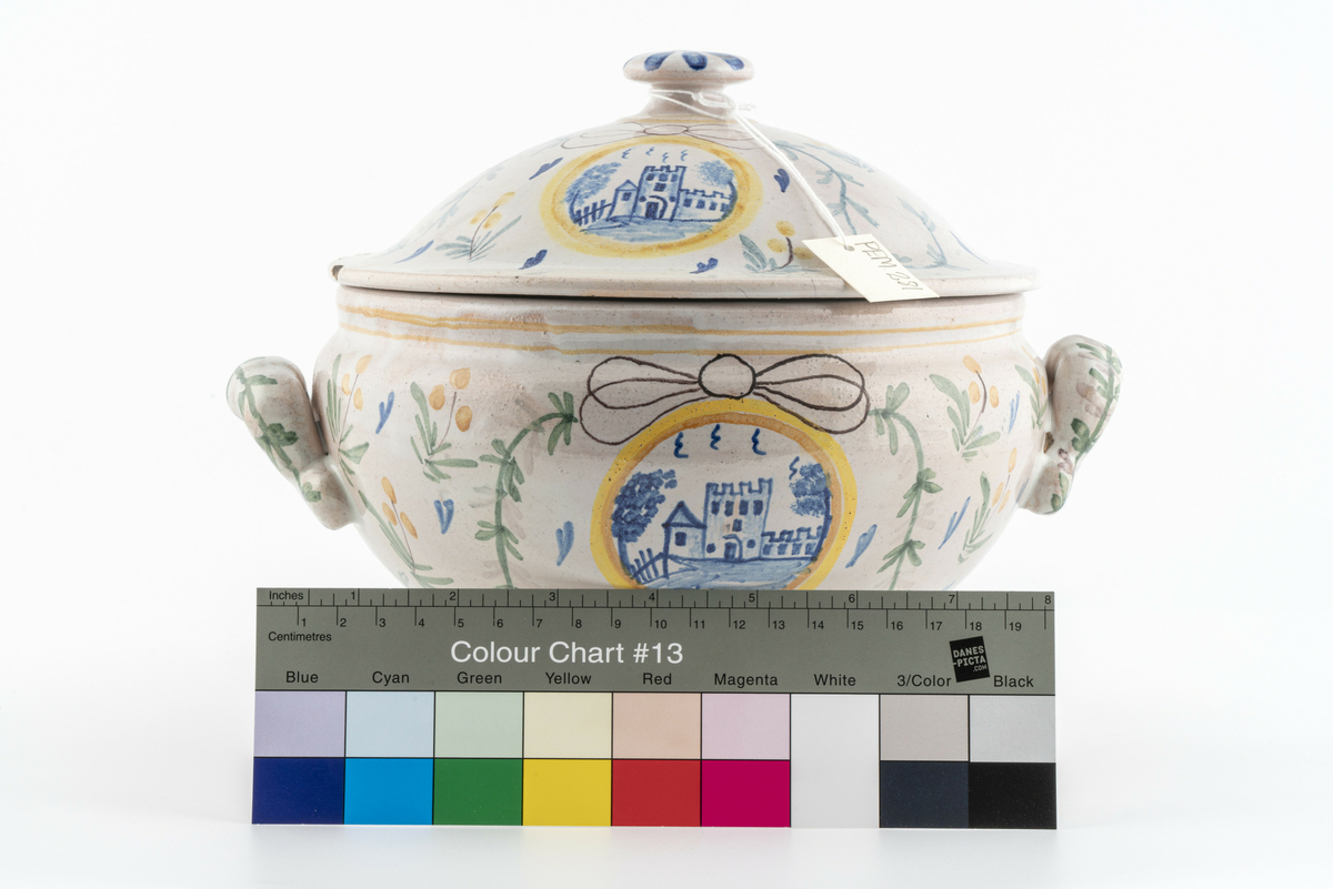 Rund terrin på stett. I keramikk, med lokk og to hanker. Beige glasur i bunn, påmalt dekor av blomster, ranker og sløyfer. Motiv av borg på ene siden, motiv av vindmølle på den andre. Disse to motivene går igjen i dekoren på lokket. Mye krakelering i glasuren inni. Har tilhørt Sara Fabricius.