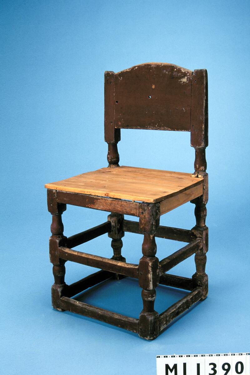 Stol av björk och barrträ målad i rödbrunt. 1600-talstyp. Balusterformade ben, nedtill och på mitten förbundna med slåar. Ryggen har balusterformade ståndare och liggande hel ryggbricka. Nytillverkad sits. Balusterformade och raka fram och bakben förenade med varandra genom tvärslåar vid bak och frambenen samt på sidorna. Fyra tvärslåar vid benens fötter och fyra i mitten. En rak sarg. Sitsen fastsatt till sargen med hjälp av träpluggar. Ryggpartiet består av balusterformade och korta ryggståndare och en liggande ryggbricka. Ryggbrickan är något avrundad vid överkanten. Stolen är rödmålad. Det finns märkning på baksargens baksida: "nes", "axd" Tillstånd: Färgen är nött. Sidosargarna och sitsen är nya.  Enligt liggaren är stolen antingen inkommen 1919 eller 1941. 

