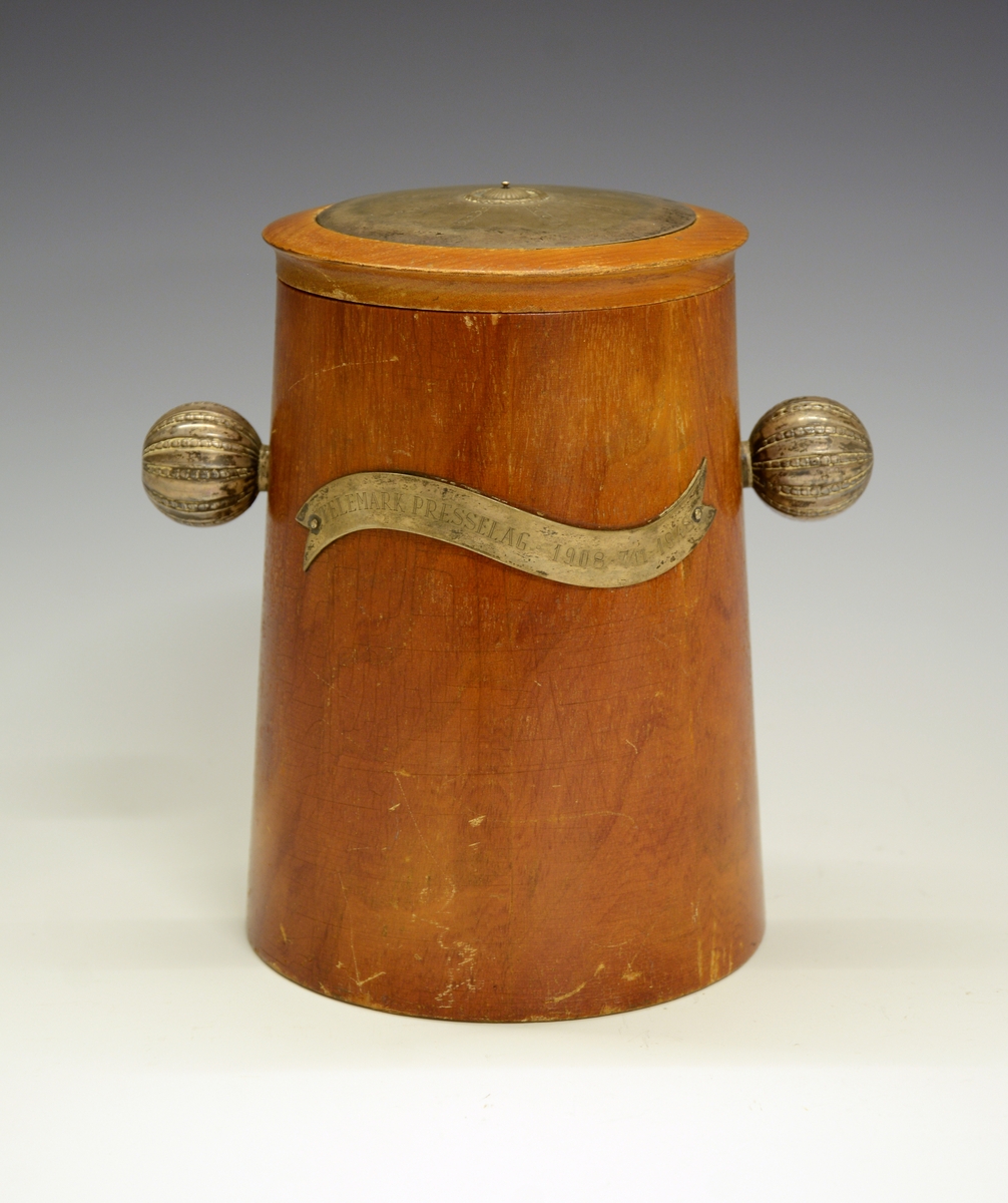 Krukke med lokk. Tre med sølvdetaljer. Sølvbånd med gravering på hver side: FRA NORSK PRESSEFORBUND og TELEMARK PRESSELAG 1908 - 7/11- 1948.