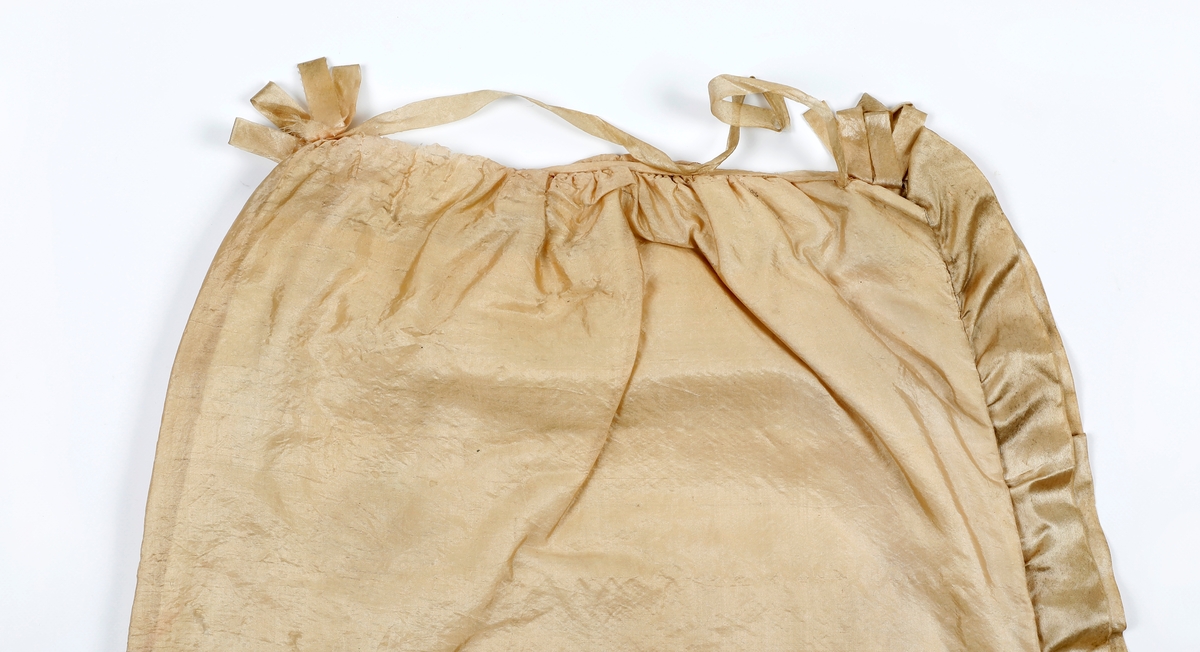 Dåpskåpe i tynn silke med rysjekappe langs alle sidene. På hodet er stoffene rynket sammen og det er laget en rosett av silkebånd foran og bak med silkebånd imellom. Kåpen er fore med bomullslerret. Det er festet silkebånd til knyting på fire steder på hver side av kåpen.Silken er skjør og hullete.