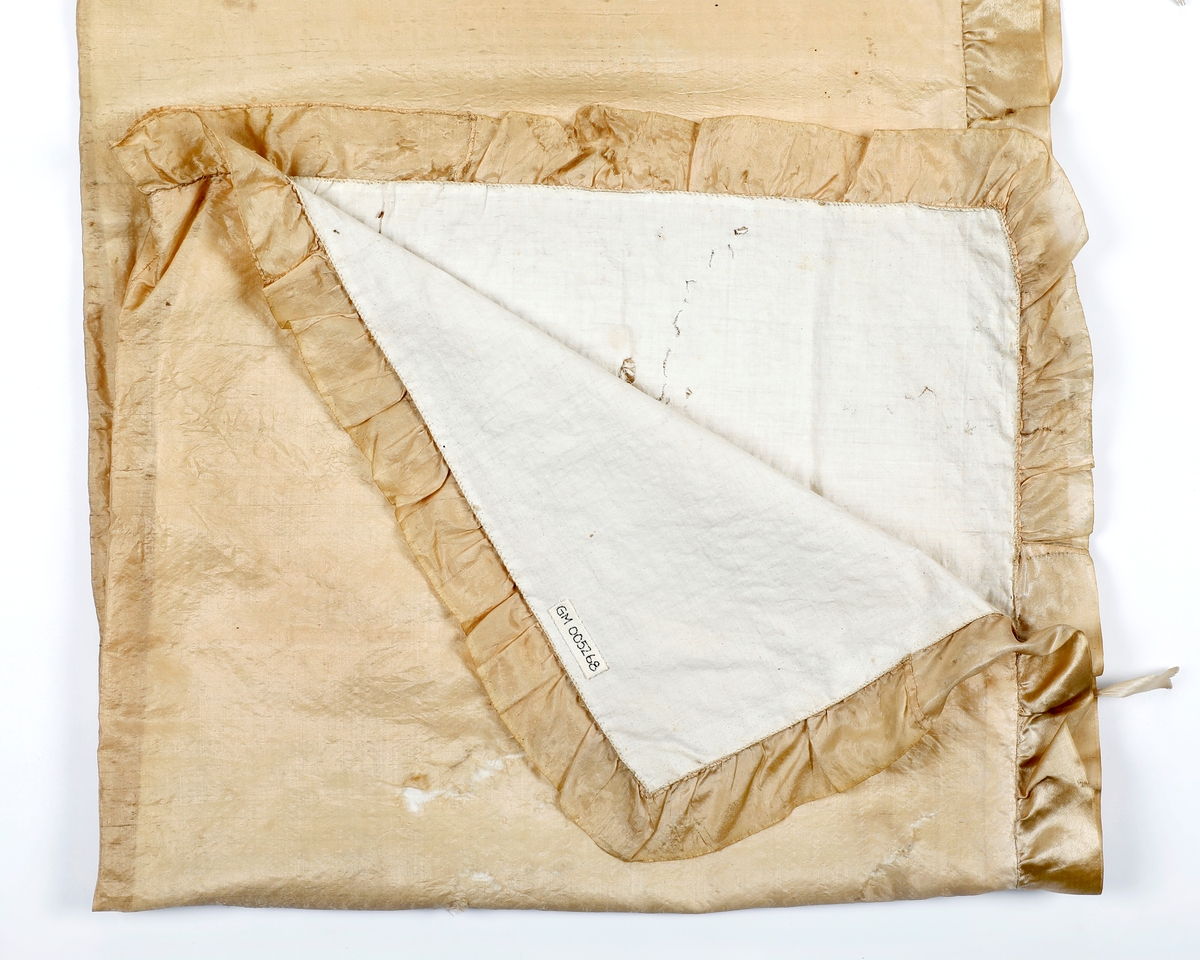 Dåpskåpe i tynn silke med rysjekappe langs alle sidene. På hodet er stoffene rynket sammen og det er laget en rosett av silkebånd foran og bak med silkebånd imellom. Kåpen er fore med bomullslerret. Det er festet silkebånd til knyting på fire steder på hver side av kåpen.Silken er skjør og hullete.