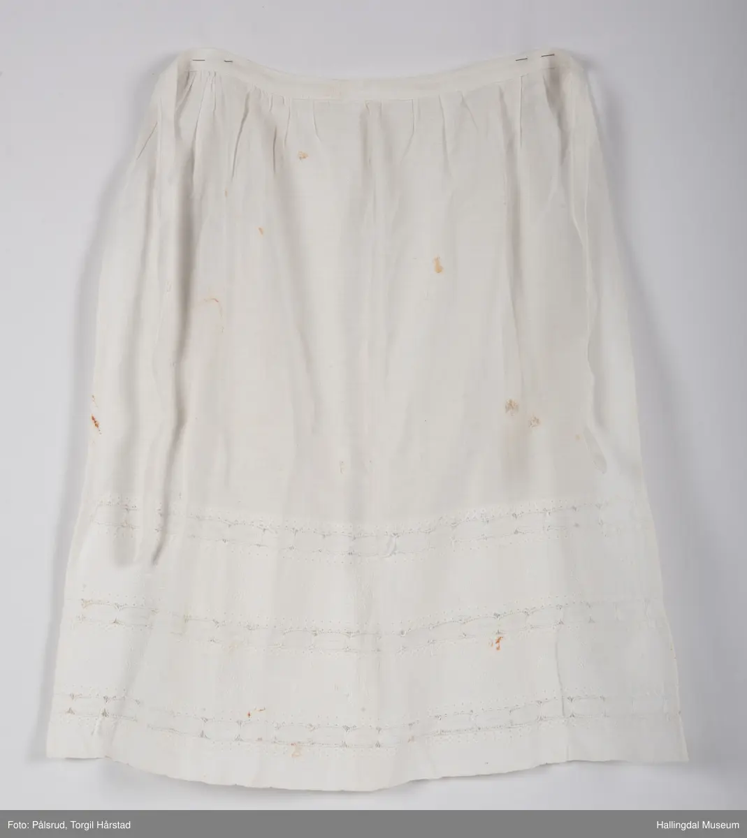 Et enkelt, hvitt forkle, mulig til serveringsformål. To bånd for å knytes sammen i rygg. Nederst på ca. en tredjedel av forkleet har stoffet blondedekor.