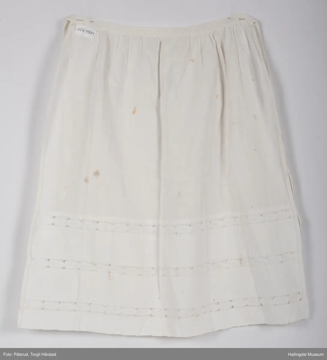 Et enkelt, hvitt forkle, mulig til serveringsformål. To bånd for å knytes sammen i rygg. Nederst på ca. en tredjedel av forkleet har stoffet blondedekor.