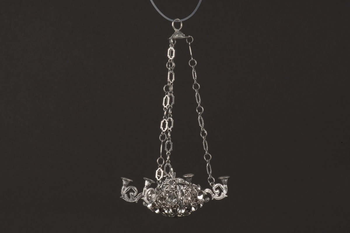 Ljuskrona till dockskåp, tillverkad av tenn.
Kronan har 6 ljusarmar, något tillböjda, och hänger i 3 kedjor.