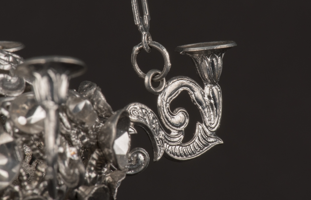 Ljuskrona till dockskåp, tillverkad av tenn.
Kronan har 6 ljusarmar, något tillböjda, och hänger i 3 kedjor.