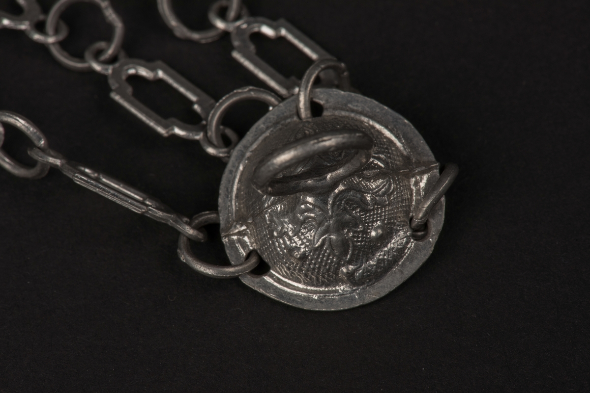 Ljuskrona till dockskåp, tillverkad av tenn.
Kronan har 6 ljusarmar, något tillböjda, och hänger i 3 kedjor.