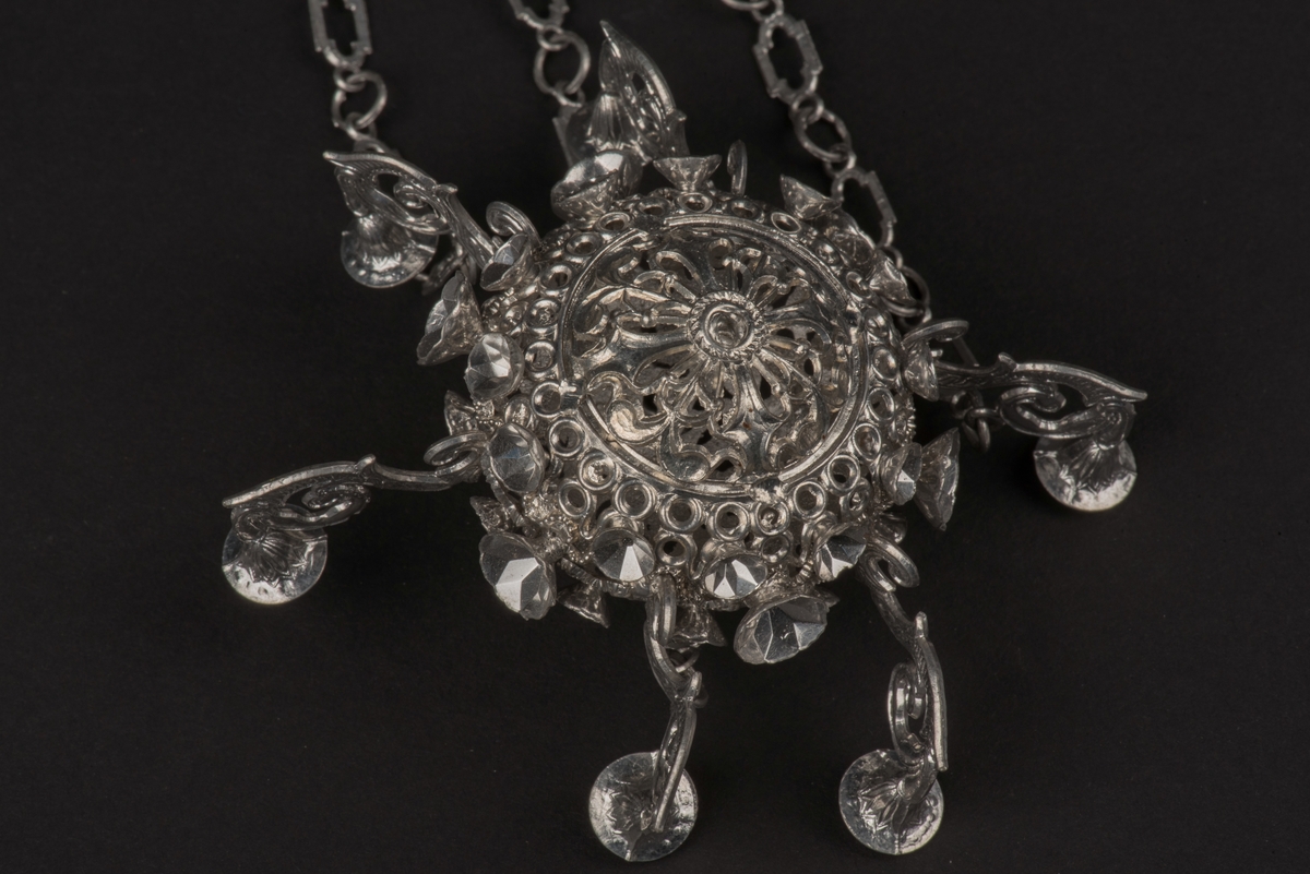 Ljuskrona till dockskåp, tillverkad av tenn.
Kronan har 6 ljusarmar, något tillböjda, och hänger i 3 kedjor.