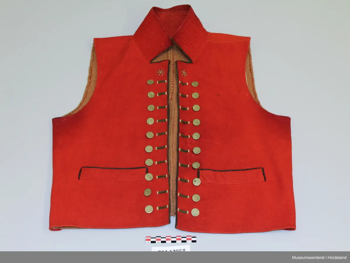 Dobbelknappa, raud mannsvest i ull med høg, raud krage. To lommer. Ei innfelt, raud stoffstripe oppå kvar skulder. Vesten er kanta med brunt silkeband framme i opninga og på lommene. Ti, tynne gullfarga knappar med knapphol på kvar side av opninga. Knapphola er sydde med ein brungrønfarga tråd, og med ein gulfarga- i endane. Lite, brodert stjerne-/blomstermotiv på kvar side oppe i spissane ved halsen, med dei same to trådfargane. Den høgreiste kragen er dobbel og er stiva av med tette horisontale saumar frå baksida. Tjukt, brunt vadmålsstoff i rygg. Framstykka er fora med bomullstoff, men det er linstoff i lommene. I spissane ved halsen er det på baksida sydd inn ein liten bit av eit mønstra bomullstoff i raudt, blått og ubleika farge. Heile vesten er handsauma.