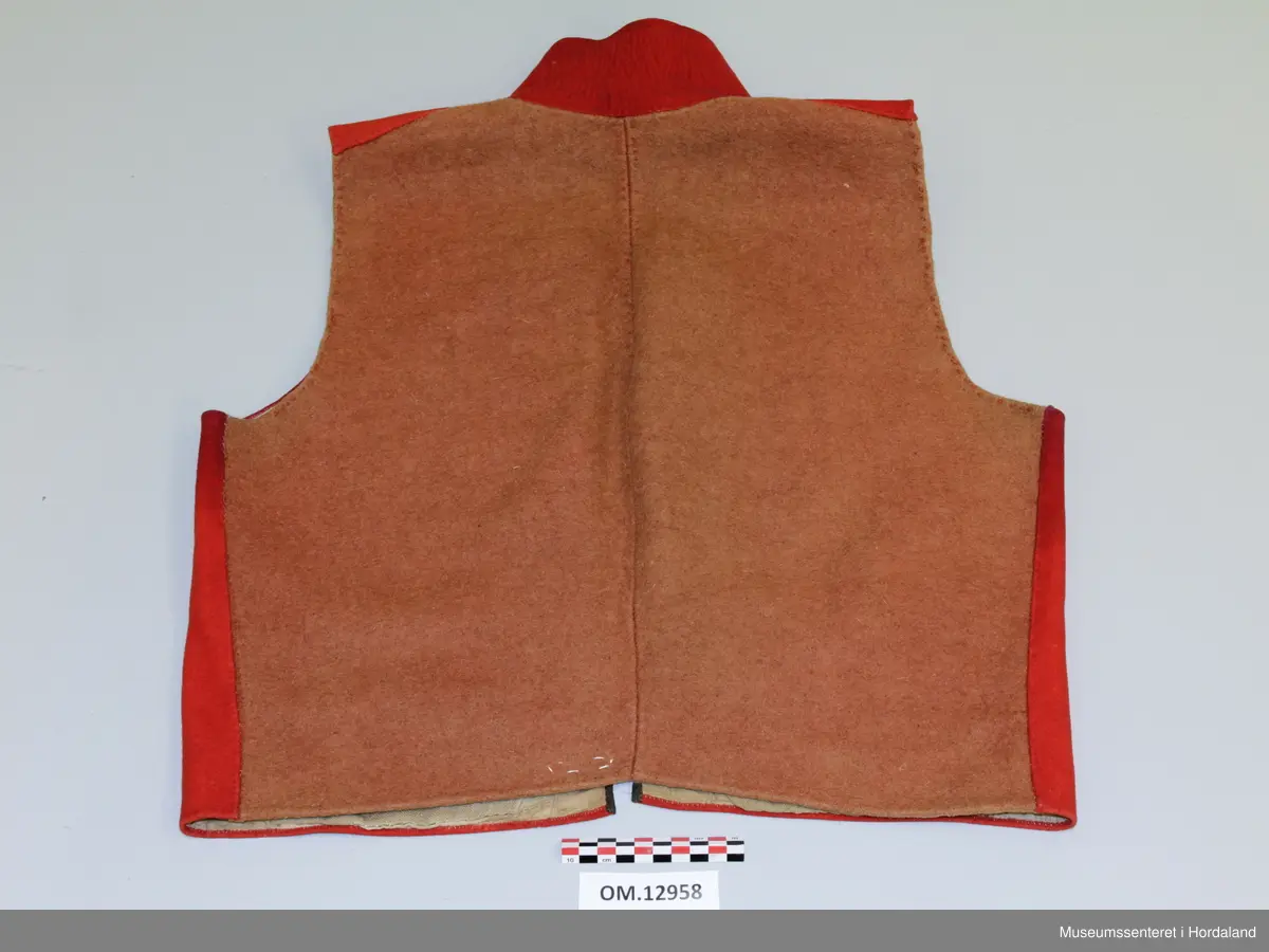 Dobbelknappa, raud mannsvest i ull med høg, raud krage. To lommer. Ei innfelt, raud stoffstripe oppå kvar skulder. Vesten er kanta med brunt silkeband framme i opninga og på lommene. Ti, tynne gullfarga knappar med knapphol på kvar side av opninga. Knapphola er sydde med ein brungrønfarga tråd, og med ein gulfarga- i endane. Lite, brodert stjerne-/blomstermotiv på kvar side oppe i spissane ved halsen, med dei same to trådfargane. Den høgreiste kragen er dobbel og er stiva av med tette horisontale saumar frå baksida. Tjukt, brunt vadmålsstoff i rygg. Framstykka er fora med bomullstoff, men det er linstoff i lommene. I spissane ved halsen er det på baksida sydd inn ein liten bit av eit mønstra bomullstoff i raudt, blått og ubleika farge. Heile vesten er handsauma.