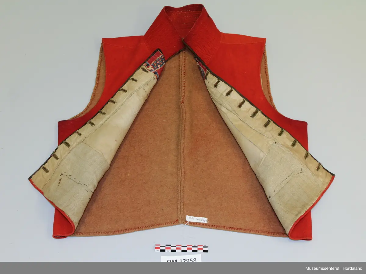 Dobbelknappa, raud mannsvest i ull med høg, raud krage. To lommer. Ei innfelt, raud stoffstripe oppå kvar skulder. Vesten er kanta med brunt silkeband framme i opninga og på lommene. Ti, tynne gullfarga knappar med knapphol på kvar side av opninga. Knapphola er sydde med ein brungrønfarga tråd, og med ein gulfarga- i endane. Lite, brodert stjerne-/blomstermotiv på kvar side oppe i spissane ved halsen, med dei same to trådfargane. Den høgreiste kragen er dobbel og er stiva av med tette horisontale saumar frå baksida. Tjukt, brunt vadmålsstoff i rygg. Framstykka er fora med bomullstoff, men det er linstoff i lommene. I spissane ved halsen er det på baksida sydd inn ein liten bit av eit mønstra bomullstoff i raudt, blått og ubleika farge. Heile vesten er handsauma.