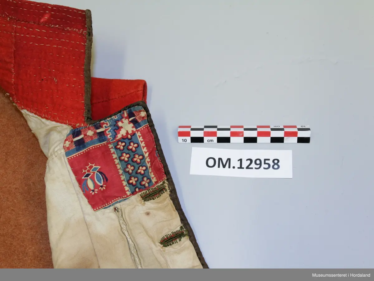 Dobbelknappa, raud mannsvest i ull med høg, raud krage. To lommer. Ei innfelt, raud stoffstripe oppå kvar skulder. Vesten er kanta med brunt silkeband framme i opninga og på lommene. Ti, tynne gullfarga knappar med knapphol på kvar side av opninga. Knapphola er sydde med ein brungrønfarga tråd, og med ein gulfarga- i endane. Lite, brodert stjerne-/blomstermotiv på kvar side oppe i spissane ved halsen, med dei same to trådfargane. Den høgreiste kragen er dobbel og er stiva av med tette horisontale saumar frå baksida. Tjukt, brunt vadmålsstoff i rygg. Framstykka er fora med bomullstoff, men det er linstoff i lommene. I spissane ved halsen er det på baksida sydd inn ein liten bit av eit mønstra bomullstoff i raudt, blått og ubleika farge. Heile vesten er handsauma.